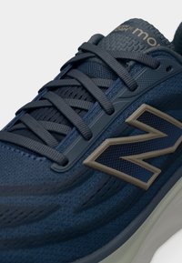 Σκούρο μπλε αθλητικό παπούτσι με υφασμάτινη υφή, σκούρα κορδόνια, μπεζ λογότυπο "N" στο πλάι και αναπαυτική σόλα σχεδιασμένη για αθλητική χρήση.