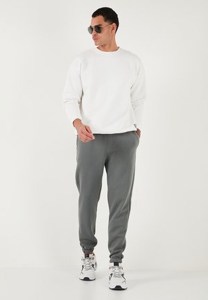 Hanorac alb cu guler rotund, asortat cu pantaloni joggers gri cu manșete elastice. Ochelari de soare negri și adidași albi completează ținuta.