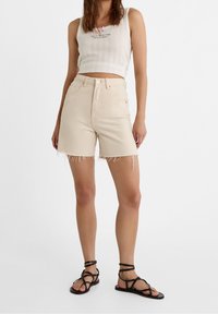 Mujer con pantalones cortos de tiro alto desgastados en color beige, top corto blanco con un pequeño lazo rosa y sandalias planas negras con tiras.