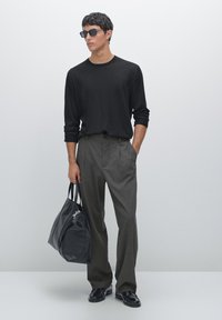 Maglietta nera a maniche lunghe, pantaloni strisciati grigi, scarpe nere e una grande borsa in pelle nera. Modello con occhiali da sole, in piedi contro uno sfondo grigio.