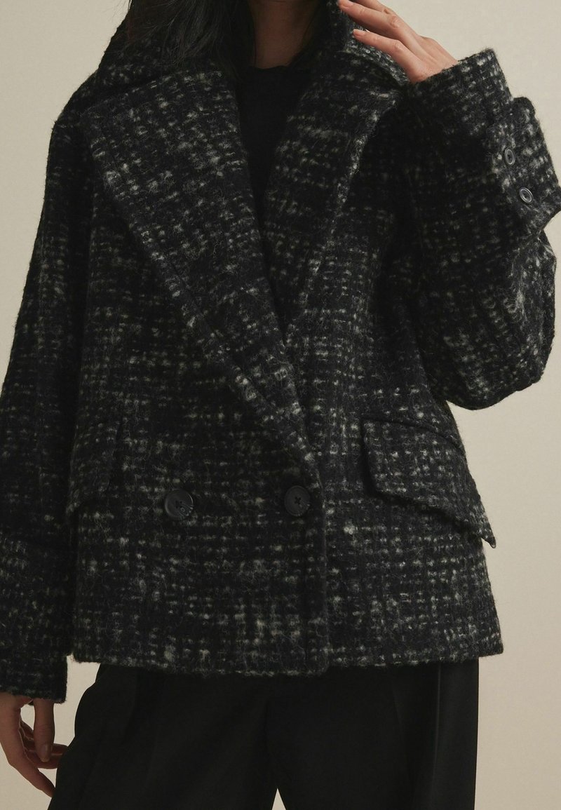 Femme portant un manteau en laine à carreaux noirs et gris, croisé, avec de larges revers et des poignets boutonnés, associé à un pantalon noir.