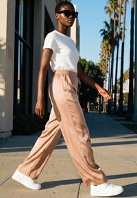 Frau mit Sonnenbrille, weißem T-Shirt, beigen, weit geschnittenen Hosen und weißen Turnschuhen, die bei Sonnenschein auf einem Bürgersteig mit Palmen im Hintergrund geht.
