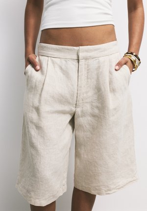 Beige linen culottes featuring a relaxed fit, pleats at the waist, and side pockets, with a textured and slightly crinkled appearance.