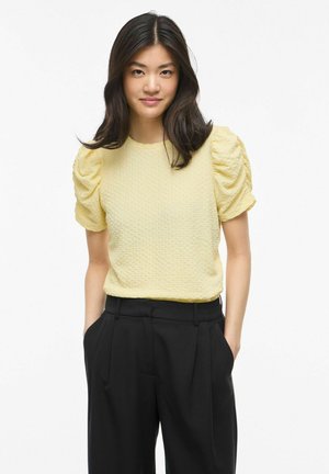 Kobieta z długimi ciemnymi włosami, ubrana w żółty teksturowany top z bufiastymi rękawami oraz wysokiej talii czarne spodnie, stojąca na białym tle, ręce w kieszeniach.