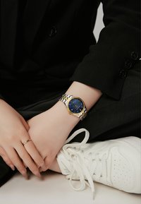 Reloj bicolor en plata y oro con un dial texturizado azul y función de fecha. Usado en una mano que descansa sobre una tela negra, cerca de unas zapatillas blancas.