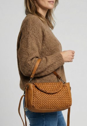 Femme portant un pull en maille marron et un jean bleu, avec un sac à main en cuir tressé beige porté à l'épaule.
