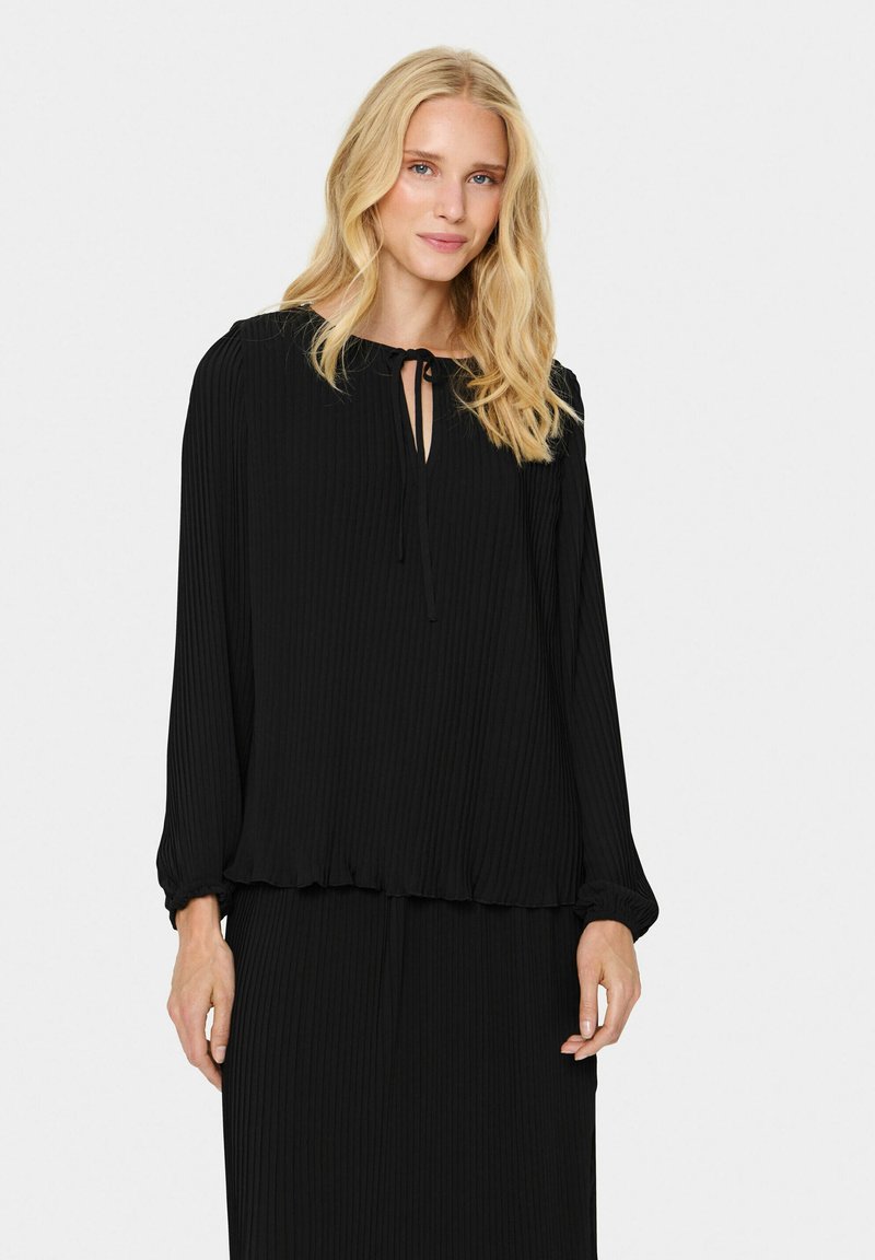 Blusa nera a maniche lunghe con texture a coste, scollo con fiocco e orlo arricciato. Il tessuto appare morbido e fluente.