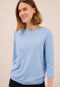 Frau mit schulterlangem, hellbraunem Haar, die ein hellblaues Langarmshirt und schwarze Hosen trägt, leicht lächelnd vor einem schlichten beigen Hintergrund.