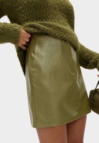 Gonna mini in pelle verde oliva con una leggera lucentezza, dotata di tasche laterali e linee pulite. Abbinata a un maglione fuzzy verde oliva a trama.