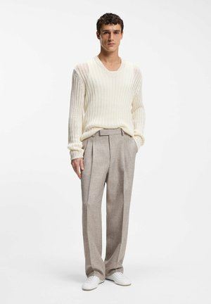 Man standing wearing a white knit sweater, beige high-waisted trousers, white sneakers, with one hand in pocket on a plain white background.