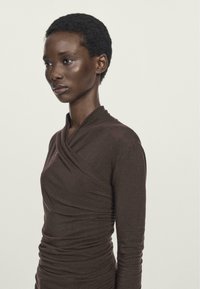 Woman with short curly hair wearing a fitted long-sleeve brown top with a wrapped neckline, looking to the left on a plain background.