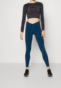 Svart croppad sweatshirt med långa ärmar och teal leggings, med en slät textur och en bred midjeband. Svarta träningsskor fulländar outfiten.