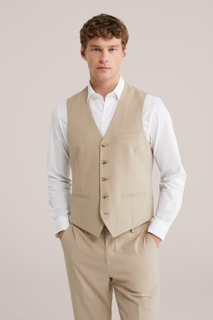 Ung mand iført beige knappet vest over hvid skjorte med matchende beige bukser, hænderne i lommerne, neutral baggrund.
