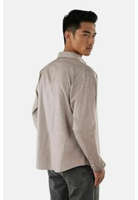 Lys beige langermet skjorte med krage, synlig søm og knapper ved mansjettene. Ledig passform, brukt sammen med mørkegrå bukser.