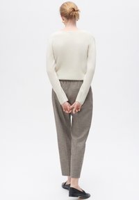 Ljus creamfärgad långärmad stickad tröja i kombination med bruna texturerade byxor. Personen vänder ryggen mot oss, händerna sammanfogade bakom ryggen, svarta skor synliga.