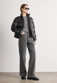 Melna pūšamā jaka ar augstu apkakli un spīdīgu virsmu, apvienota ar svītrainu garām piedurknēm krekliņu un pelēkiem platām kājām džinsiem, pabeigta ar melnām zābakiem.