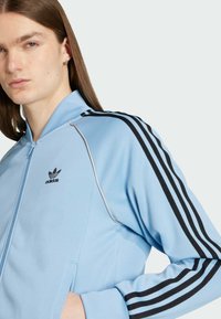 Světle modrá zipová bunda vyrobená z hladké tkaniny. Má černé pruhy na rukávech a vyšívané černé logo Adidas na hrudi.