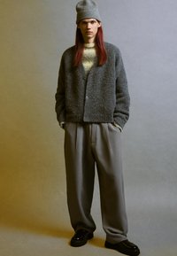 Cardigan grigio oversize lavorato a maglia con scollatura a V e bottoni, abbinato a pantaloni grigi larghi e scarpe nere. Presenta un berretto grigio.