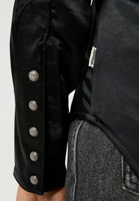 Czarna satynowa koszula z długim rękawem ma teksturowane wykończenie, guziki na mankietach oraz kontrastowy materiał na dole z metką marki.