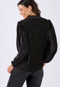 Blusa nera a costine con dettagli in oro metallico. Maniche a sbuffo e scollatura arricciata. Indossata con pantaloni neri lucidi.