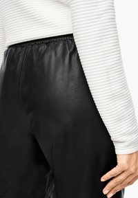 Pantalon en cuir noir avec une texture lisse et une taille élastique, associé à un haut à manches longues rayé blanc, laissant entrevoir une partie de l'articulation.