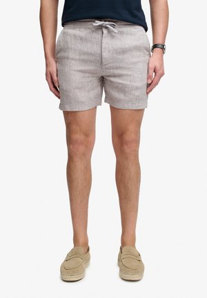 Mężczyzna ubrany w beżowe lniane szorty ze ściągaczem, granatową koszulę, zegarek na nadgarstku i beżowe wsuwane buty, stoi na tle białego tła.