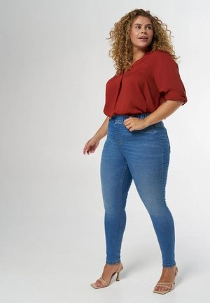 Blouse rouge à manches courtes associée à un jean skinny bleu ; texture en denim lisse ; délavage clair ; talons à brides de cheville ; mannequin posé de profil.