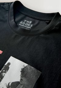 Camiseta de algodón negra con cuello redondo y estampado gráfico en tonos de gris en la parte frontal. Etiqueta "THE KOOKS" impresa en el interior del cuello.
