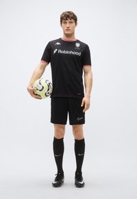 Maillot de football noir avec des accents rouges, arborant le logo « Robinhood », accompagné d'un short noir, de chaussettes jusqu'aux genoux et de chaussures de football noires. Tenant un ballon multicolore.