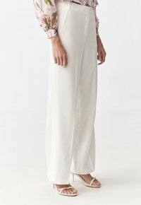 Weiße, weit geschnittene Hosen aus glattem Stoff, mit einem maßgeschneiderten Bund und seitlichen Taschen. Kombiniert mit hellen, hohen Sandalen.