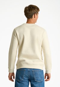 Sudadera de color crema con mangas largas, cuello redondo y puños acanalados; combinada con jeans de mezclilla azul que cuentan con dos bolsillos traseros.