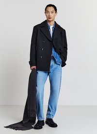 Manteau noir en laine à double boutonnage, chemise en denim bleu, jean bleu clair et bottes noires. Tenant une écharpe gris foncé à franges.