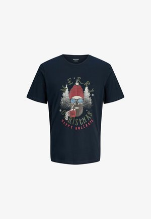 Un garçon se tenant debout, portant des chaussures beiges, un jean large délavé, un t-shirt graphique bleu marine et une chemise à carreaux ouverte, regardant vers la gauche.