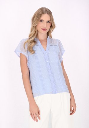 Blusa blu chiaro con maniche corte in voile, texture a pois, chiusura con bottoni, dettagli plissettati e scollatura arricciata.