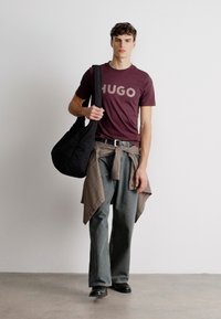 Burgundi T-särk "HUGO" graafikuga, hallid ülepaisutatud püksid, ruudulise särgi selja ümber seotud, must kott, mustad jalanõud, sile tekstuur, lõdva disainiga.