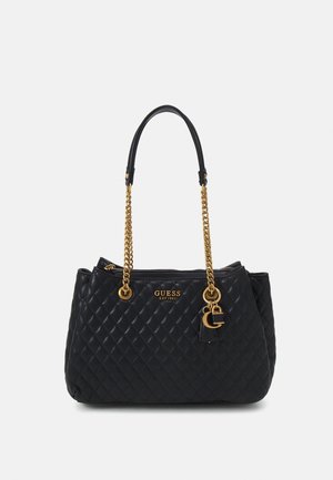 Schwarze, gesteppte Lederhandtasche mit goldenen Kettengriffen, goldenem "GUESS"-Logo und dekorativem goldenen Anhänger vor weißem Hintergrund.