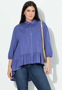 Blusa viola con bottoni, design plissettato, maniche a tre quarti, orlo asimmetrico e texture di tessuto liscia, indossata con jeans scuri.