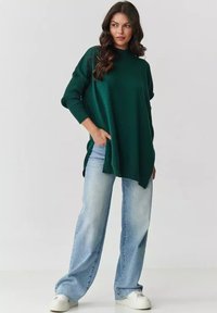 Ciemnozielony oversize'owy sweter z ribbowanymi rękawami. Połączony z jasnoniebieskimi dżinsami o szerokich nogawkach i białymi trampkami. Gładka tekstura, luźny krój.