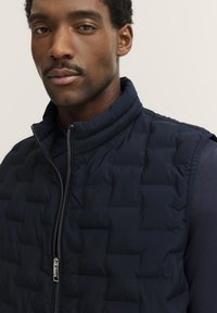 Marineblauer gesteppter Weste mit hohem Kragen und silbernem Reißverschluss. Glatte Textur mit gepolsterten Quadraten, über einem dunklen Langarmshirt geschichtet.