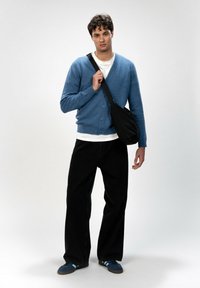 Blauer Strickcardigan, weißes T-Shirt, schwarze weite Hose und blaue Turnschuhe. Schwarze Umhängetasche mit minimalistischem Design, vor einem hellen Hintergrund stehend.