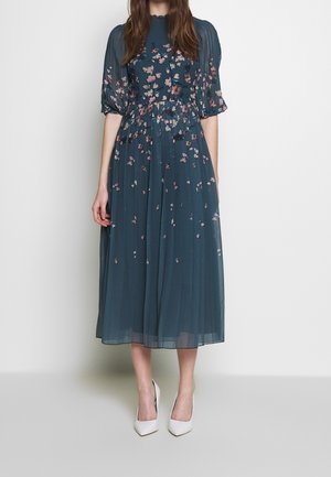 Kvinde iført en teal midi kjole med pink blomsterprint, pufærmer og hvide spidse høje hæle, stående op ad en ensfarvet væg.