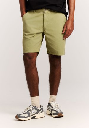 Man die olijfgroene korte broek, crèmekleurige sokken en grijs-witte sportschoenen draagt, staand tegen een effen achtergrond.