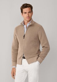 Beige geribbelde trui met ritssluiting, opstaande kraag en twee zakken, gecombineerd met een lichtblauw gestreept overhemd en witte broek. Soepele textuur.