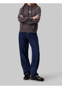 Felpa grigia con logo frontale, polsini a costine, abbinata a jeans a gamba larga blu scuro e scarpe chunk nere. Tessuto liscio e vestibilità rilassata.