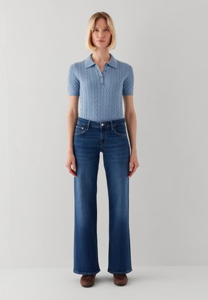 Frau, die ein hellblaues, kurzärmeliges Strick-Poloshirt und dunkelblaue ausgestellte Jeans mit braunen Schuhen vor einem einfarbigen hellgrauen Hintergrund trägt.