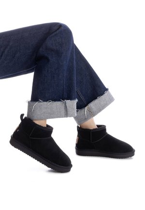Botas de tobillo de ante negro con un suave forro interior, perfil bajo y suela texturizada, combinadas con jeans oscuros enrollados.