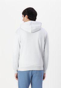 Sudadera gris con cordón, puños y dobladillo acanalados. Tela suave y corte relajado. Llevada con pantalones jogger de color azul claro, vista desde la parte de atrás.