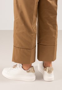 Zapatillas blancas con un acento dorado texturizado en el talón, que presentan una puntera redonda, suela blanca gruesa y detalles decorativos cerca de los cordones.