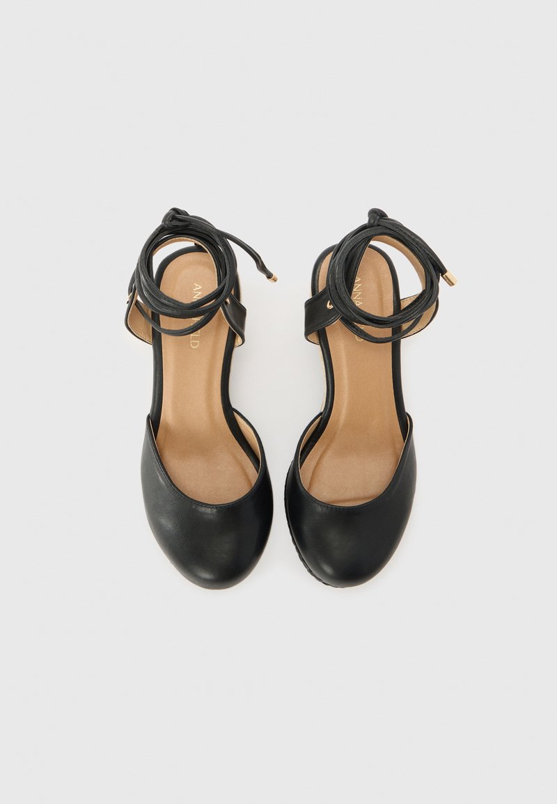 Sandales à talons hauts en cuir noir avec des brides de cheville, bouts ronds et semelles intérieures beiges. Le détail des brides présente des accents dorés pour un intérêt supplémentaire.