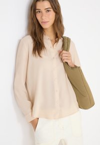 Jeune femme aux longs cheveux bruns portant un chemisier beige à boutons et un pantalon blanc, tenant un sac matelassé vert olive sur une épaule.